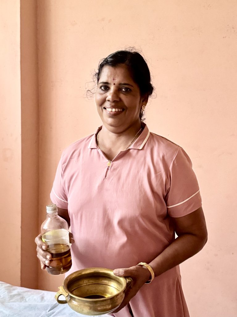 Sarojam, Massage Therapist at Prakruthi Ayurveda Cherai Beach, holds a bottle of Ayurvedic oil and a brass massage vessel